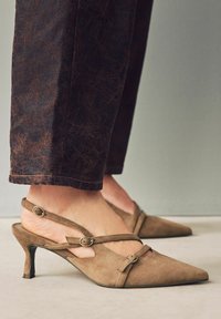 Ruskean mokkanahkaiset slingback-kengät, joissa on terävä kärki, hihna ja solkiyksityiskohdat sekä 2 tuuman stiletto-korko. Yhdistetty tummiin teksturoituihin housuihin.