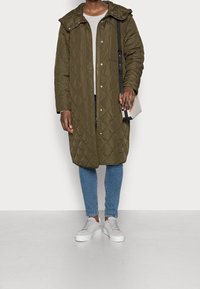 Cappotto lungo in verde oliva trapuntato con chiusura a zip e bottoni, cappuccio e tasche laterali, abbinato a jeans azzurri chiari e sneaker bianche.