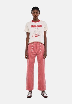 Femme portant un t-shirt blanc avec le texte rouge « Main Sail » et un graphique de voilier, assorti à un pantalon taille haute rayé rouge et blanc ainsi qu'à des mocassins.