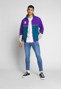 Chaqueta colorida con paneles morados, azul verdoso y beige; cuenta con un cuello y bolsillos con cremallera. Llevada sobre una camisa clara y pantalones vaqueros azules.