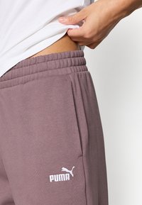 Pánske tepláky Puma v tlmenom marhuľovom odtieni, s elastickým pásom, vreckami a bielou výšivkou loga Puma na stehne.