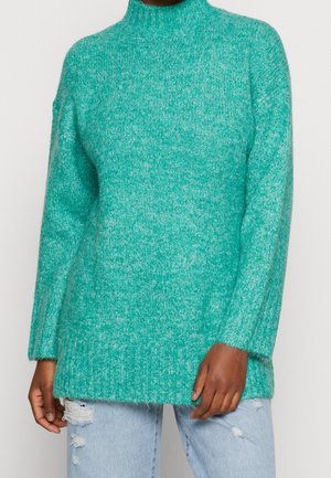 Pull en maille duveteuse vert sarcelle avec col montant porté avec un jean bleu clair déchiré sur une personne devant un fond uni.