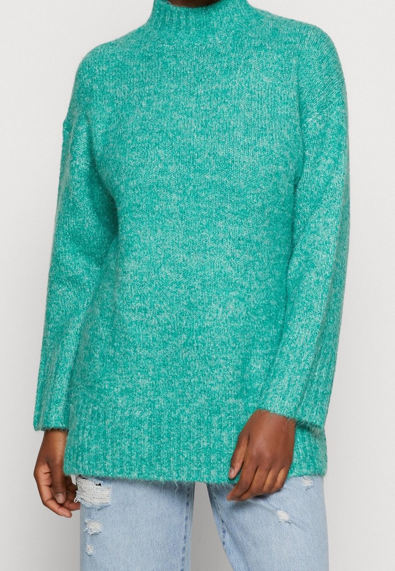 Pull en maille duveteuse vert sarcelle avec col montant porté avec un jean bleu clair déchiré sur une personne devant un fond uni.