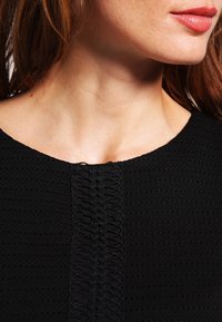 Svart texturerad plagg med ett vävt mönster längs framsidan. Urringningen är rundad med en subtil, ren finish.