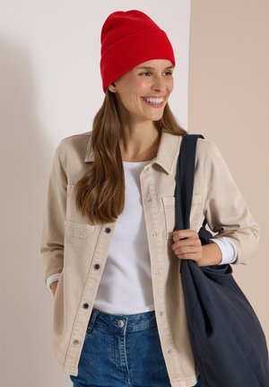 Kvinde smilende iført en rød hue, beige jakke, hvid skjorte, blå jeans og bærende en mørkeblå skuldertaske.