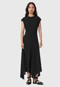 Žena v černých šatech bez rukávů s asymetrickým lemem a černými plochými botami se širokými kotníkovými pásky a sponami, stojící na šedém pozadí.