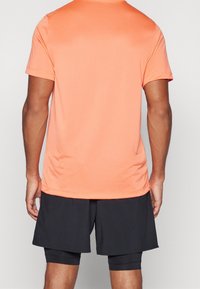 Kortærmet sportstrøje med rund hals i lyst koral, lavet af letvægtsstof; kombineret med sorte atletikshorts. Glat tekstur.