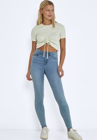 Top corto verde claro con fruncido en la parte delantera y detalle de cordón, combinado con jeans skinny de cintura alta en un azul claro y zapatillas de color blanco roto.