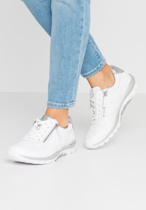 Person iført lyseblå jeans og hvide sneakers med sølv accenter og sidelommer med lynlås, stående på en lysegrå gulv.