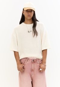 Pulover crem supradimensionat din tricot cu mâneci scurte, asortat cu pantaloni din denim roz deschis. Accesoriile includ un colier și un inel din argint, precum și o pălărie bej.