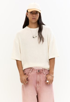 Overdimensioneret cremet strikket sweater med korte ærmer, parret med lyserøde denim bukser. Accessories inkluderer en sølvhalskæde og ring, samt en beige kasket.