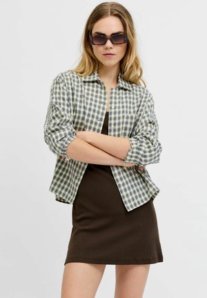 Kvinne med mørke solbriller, en grønn- og hvit rutete skjorte over en mørkebrun kjole, står med armene krysset foran en ensfarget bakgrunn.