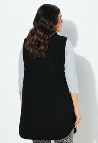 Nainen, jolla on laineileva ruskea hius, pukeutuu mustaan teksturoituun hihattomaan liiviin vaaleanharmaan paidan ja leopardikuvioisten housujen päällä, seisoo sivuttain valkoista seinää vasten.