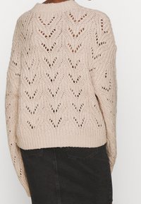 Beige gebreide trui met een chevronpatroon, opengewerkt ontwerp en een geribde hals. De mouwen zijn breed en de trui loopt taps toe bij de zoom.
