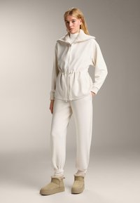 Biela mikina na zips s vysokým golierom a sťahovacím pásom, v kombinácii s rovnakými jogger nohavicami a béžovými členkovými topánkami so zateplením.