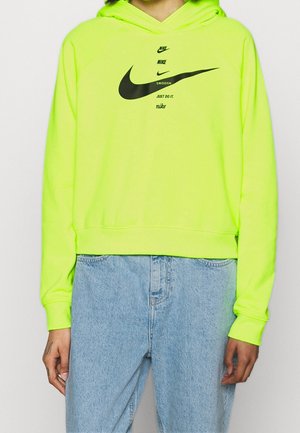 Person trägt einen neongelben Nike-Hoodie mit schwarzem Swoosh und Schriftzug, kombiniert mit hellblauen Jeans, steht vor einem einfarbigen Hintergrund.