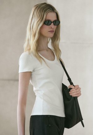 Donna con capelli biondi lunghi che indossa occhiali da sole neri, top bianco a maniche corte, pantaloni neri, e tiene una borsa nera a tracolla.