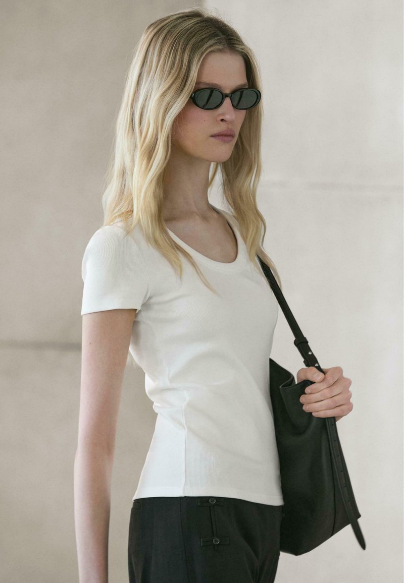 Frau mit langen blonden Haaren, die schwarze Sonnenbrille, ein weißes kurzärmeliges Oberteil und schwarze Hose trägt und eine schwarze Umhängetasche hält.