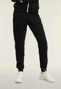 Una persona wearing pantaloni jogging neri e sneakers bianche contro uno sfondo chiaro e uniforme, in piedi con le gambe leggermente divaricate.
