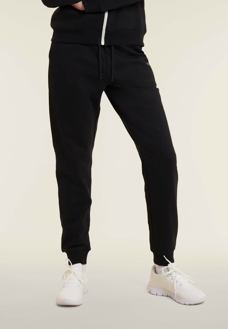 Una persona wearing pantaloni jogging neri e sneakers bianche contro uno sfondo chiaro e uniforme, in piedi con le gambe leggermente divaricate.