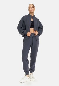 Graue Zip-Jacke mit hohem Kragen und lockerer Passform, kombiniert mit passenden, schmal zulaufenden Sweatpants. Beide Teile haben eine weiche Textur und minimales Design.