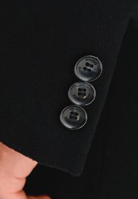 Czarna materiałowa rękaw kurtki z trzema okrągłymi guzikami. Guziki są wykonane z przejrzystego plastiku z wygrawerowanym tekstem oraz trzema dziurkami na guziki.