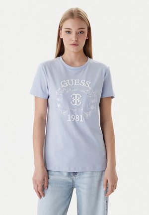 Giovane donna che indossa una t-shirt Guess 1981 azzurro chiaro e jeans chiari, in piedi davanti a uno sfondo bianco semplice.