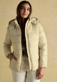 Bežová prešívaná bunda s kapucňou, so zipsom a cvokmi. Má jemný lesk a rebrované manžety. Nosená cez vzorovaný sveter.