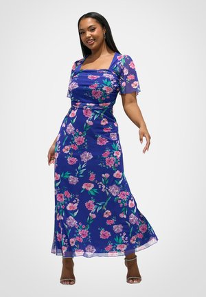 Kvinde iført en blå blomsterprint midi-kjole med korte ærmer og firkantet halsudskæring, poserer med et smil og iført sandaler med hæl.