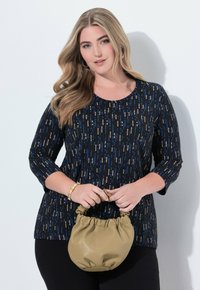 Frau mit langen blonden Haaren hält eine kleine beige geraffte Handtasche und trägt ein schwarzes Oberteil mit blauen und beigen vertikalen Punkten sowie schwarze Hosen.