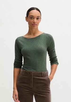 Femme aux cheveux foncés en chignon bas portant un haut vert à manches longues et un pantalon en velours côtelé marron taille haute, debout avec une main dans la poche.
