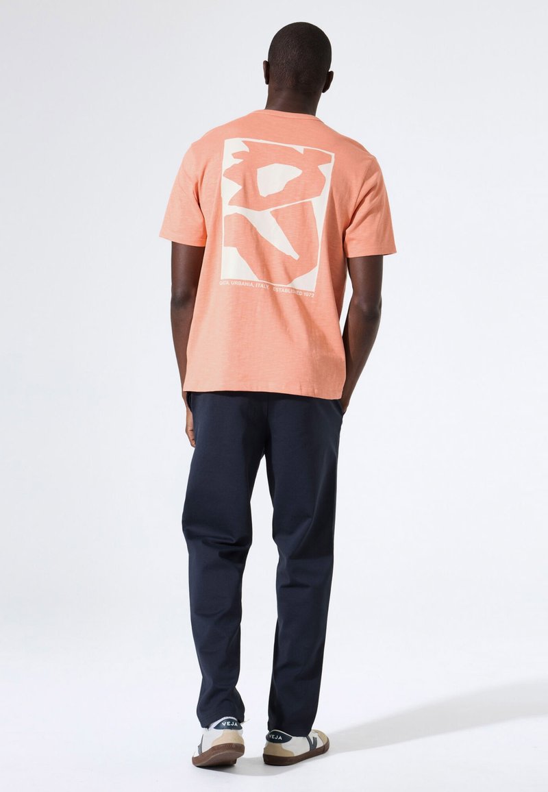 Koraalrood T-shirt met korte mouwen en een witte grafische print op de achterkant, gecombineerd met marineblauwe broek en sandalen met een wit-bruine print.