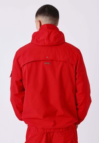 Man met kort haar in een felrode hoodie en bijpassende broek, weergegeven van achteren tegen een eenvoudige achtergrond.