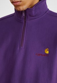 Lilla fleece-pullover med høj krave, kvart-zip åbning og rødt broderet "Carhartt" logo med gul accent. Blødt tekstur.