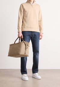 Sudadera cremada con cremallera, jeans azules y zapatillas blancas, sosteniendo un bolso de lona estructurado en color beige con acentos y asas de cuero marrón.
