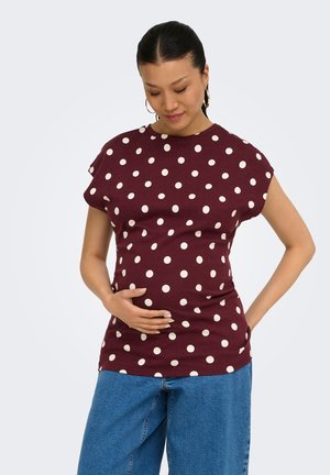 Vrouw met zwart haar in een knot, gekleed in een bordeauxrood shirt met stippen en een blauwe spijkerbroek, met de ene hand haar buik vasthoudend en de andere op haar heup.