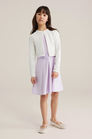 Noor tüdruk kannab valget kootud kardigani üle helelilla voltidega kleidi ja hõbedaste sädelevate Mary Jane kingade, seistes hele taustaga.