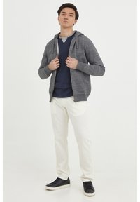 Sudadera gris con cremallera sobre una camiseta azul marino, pantalones blancos y zapatillas negras. La sudadera tiene cordones y un corte casual.