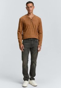 Brun langærmet henley-shirt med to sorte knapper, båret med mørkegrå jeans og hvide sneakers. Enkel, tekstureret stofdesign.