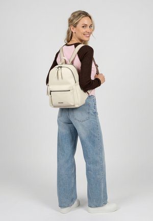 Femme souriante portant un t-shirt à manches longues rose et marron, un jean bleu clair, des baskets blanches et un sac à dos beige sur un fond uni.