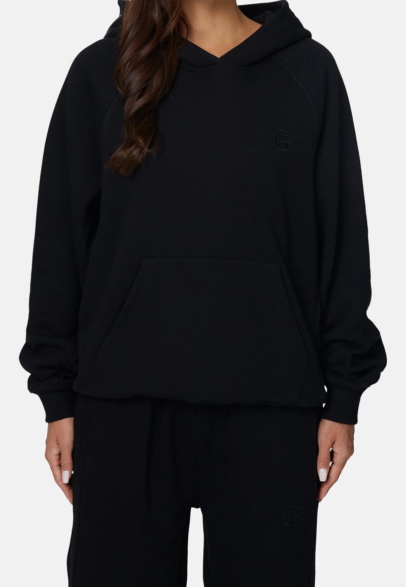Femme portant un sweat à capuche noir oversize avec poche kangourou et pantalon noir assorti sur fond uni.