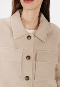 Kvinde iført en beige uldjakke med store knapper og to forreste klaplommer, vist fra brystet og op.