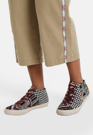 Jambes portant un pantalon beige raccourci avec des bandes latérales à motifs et des baskets multicolores en patchwork avec des lacets rouges sur fond blanc.