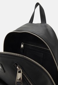 Sac à dos en cuir noir avec fermeture éclair argentée, comprenant un compartiment intérieur et une petite poche zippée. Texture lisse, design minimaliste.