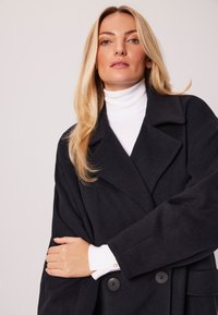 Czarny, dopasowany płaszcz z wyłogi, podwójnym zapięciem i miękkim, teksturowanym materiałem, nałożony na biały golf.