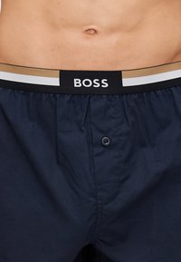 Navy blue boxer shorts with a contrasting black waistband featuring white and gold stripes and a BOSS logo. Equipped with a button closure.