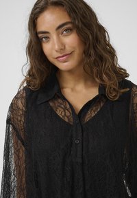 Blouse en dentelle noire à manches longues, avec un devant à boutons, un col et un motif de dentelle complexe, offrant une texture transparente.