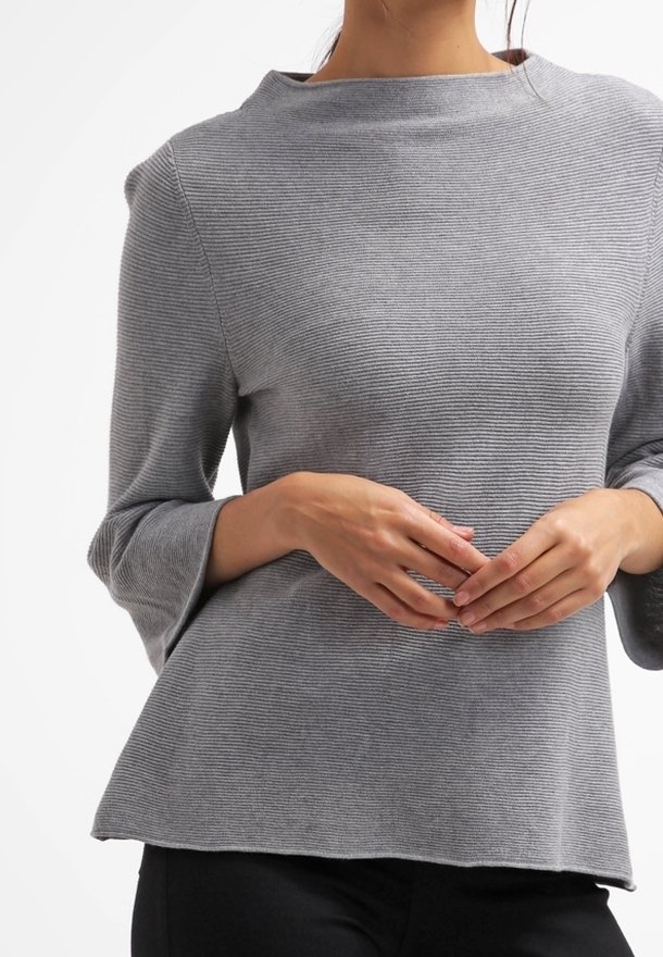 Femme portant un haut gris côtelé à manches longues légèrement évasées, debout, les mains jointes devant elle de manière relâchée.