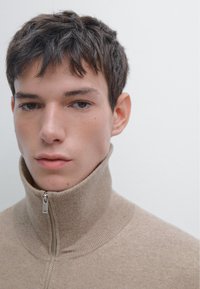 Ung man med kort mörkt hår som bär en beige zip-up-tröja med kragen helt zipad mot en enkel ljus bakgrund.