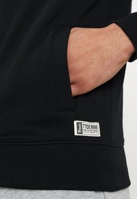 Felpa nera con tasca laterale e orlo a costine. Include un'etichetta bianca con "TIDENIM" e dettagli del marchio. Tessuto morbido.